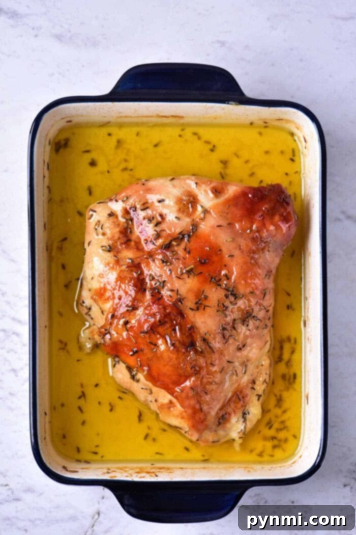 Mastering Turkey Breast Confit 5 A cooked piece of turkey breast, prepared as Turkey Confit, is shown in a rectangular blue and white baking dish filled with golden liquid, likely broth or melted butter. The turkey is seasoned with herbs. The dish is placed on a light-textured surface.