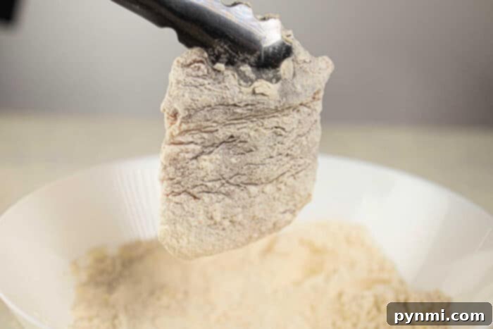 Ultimate Crispy Pork Chops 4 A piece of breaded boneless pork chops, soon to become a crispy delight, is held by tongs over a bowl of flour, ready to transform into savory deep-fried pork chops.