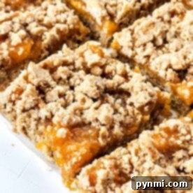 Close-up of peach bars with a crumbly topping, arranged in a grid pattern on a white surface, capturing the sweet charm of summer in each bite.