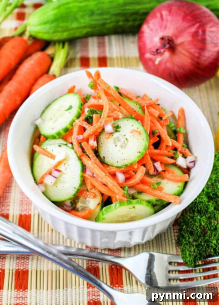 Refreshing Carrot Cucumber Salad 2 A white bowl filled with a Carrot and Cucumber Salad, featuring sliced cucumbers, julienned carrots, and chopped red onions, garnished with herbs. The bowl sits on a woven mat, surrounded by whole carrots, a cucumber, a red onion, and parsley. A fork and knife rest beside the bowl.