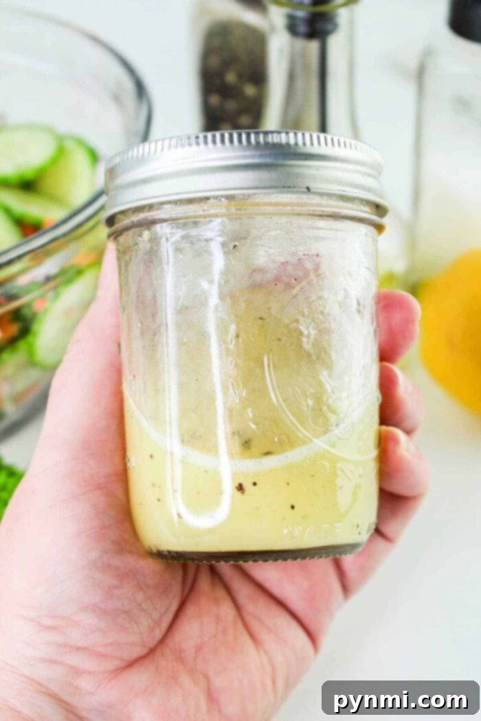 Refreshing Carrot Cucumber Salad 4 A hand is holding a glass jar filled halfway with light-colored salad dressing, perfect for a refreshing Carrot and Cucumber Salad. The jar has a metal screw-top lid. In the background, there is a bowl of sliced cucumbers, a pepper grinder, a bottle, and a lemon.