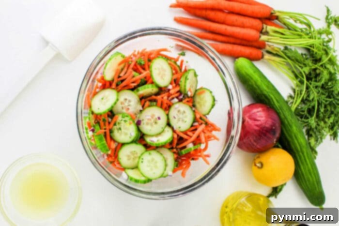 Refreshing Carrot Cucumber Salad 5 A glass bowl filled with a Carrot and Cucumber Salad, featuring sliced cucumbers, shredded carrots, and chopped herbs. Surrounding the bowl are whole carrots with greens, a cucumber, a red onion, a lemon, a small bowl of dressing, a bottle of oil, and a white kitchen tool.