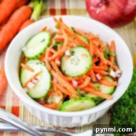 Refreshing Carrot Cucumber Salad 6 A white bowl filled with a Carrot and Cucumber Salad, featuring sliced cucumbers, julienned carrots, and chopped red onions, garnished with herbs. The bowl sits on a woven mat, surrounded by whole carrots, a cucumber, a red onion, and parsley. A fork and knife rest beside the bowl.