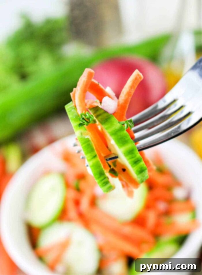 Refreshing Carrot Cucumber Salad 7 A fork holds a bite-sized portion of Carrot and Cucumber Salad, featuring cucumber slices, carrot strips, and chopped red onions. In the background, there is a bowl of similar salad ingredients and some vegetables.
