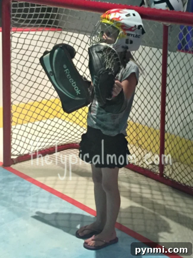 Kids engage in active learning at Discovery Cube OC's hockey exhibit, a highlight for families in Orange County. A young child in hockey gear, including a helmet, glove, and blocker, stands ready in front of a goal net. Despite wearing casual clothes and flip-flops, their focused stance embodies the spirit of interactive sports exhibits found at the Discovery Science Center Santa Ana, promoting active play and learning.