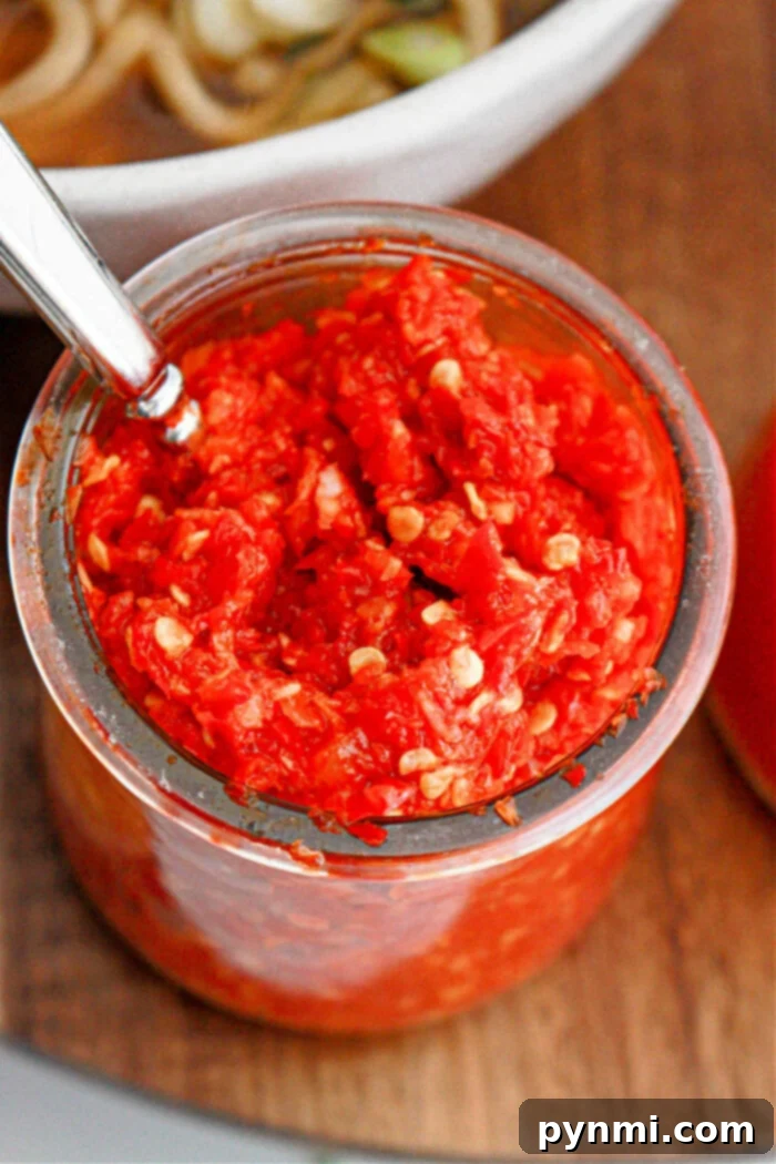 Fiery Garlic Chili Paste 6 A clear glass jar filled with chunky homemade garlic chili paste, showing visible seeds and a metal spoon inserted into the jar. It sits on a wooden surface with part of a white bowl containing food visible in the background, suggesting its use as a vibrant condiment.