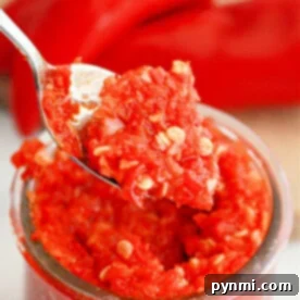 A close-up of a spoon holding a portion of vibrant red homemade garlic chili paste with visible seeds, positioned above a glass jar filled with the same rich mixture. In the soft background, whole red peppers are slightly out of focus, evoking the fresh ingredients of this savory recipe.