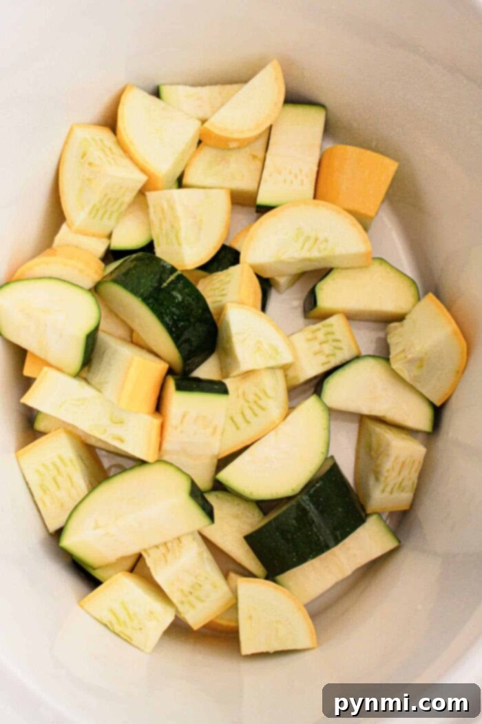 The image shows chopped pieces of zucchini and yellow squash inside a white container. The vegetables, perfect for a slow cooker recipe, are cut into uniform chunks, displaying their green and yellow peels.