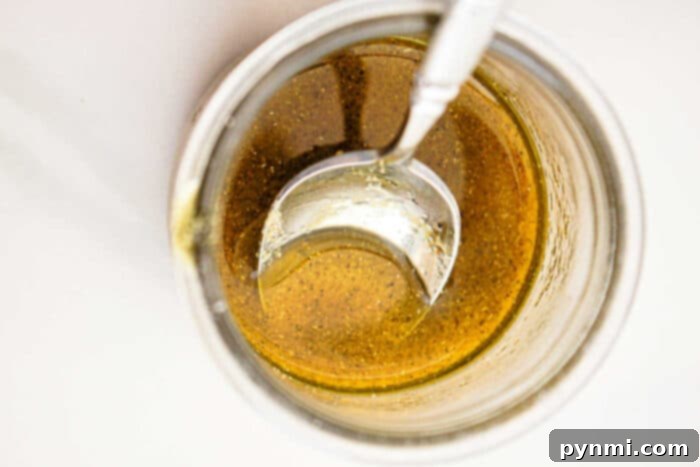 Close-up view of a glass jar filled with a golden-brown liquid, likely a sauce or dressing perfect for slow cooker vegetables, with a metal spoon partially submerged in it. The liquid has some visible seasoning or spices. The background is white and minimal.