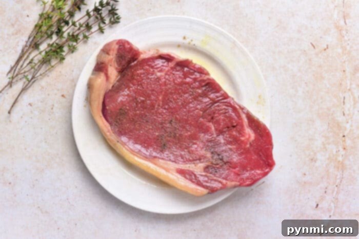 Raw beef strip loin steak seasoned with salt and pepper, placed on a pristine white plate, accompanied by fresh sprigs of thyme. This visual illustrates the initial preparation stage before cooking, emphasizing simple yet effective seasoning for optimal flavor.