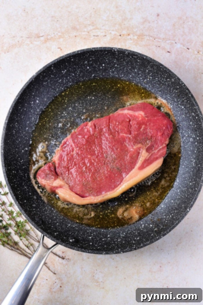 A raw strip loin steak resting in a frying pan with a small amount of oil, ready for searing. Fresh thyme sprigs lie beside the pan on a light countertop, indicating the inclusion of aromatics during the cooking process. This image captures the initial moments of preparing a beef strip loin steak on the stove.