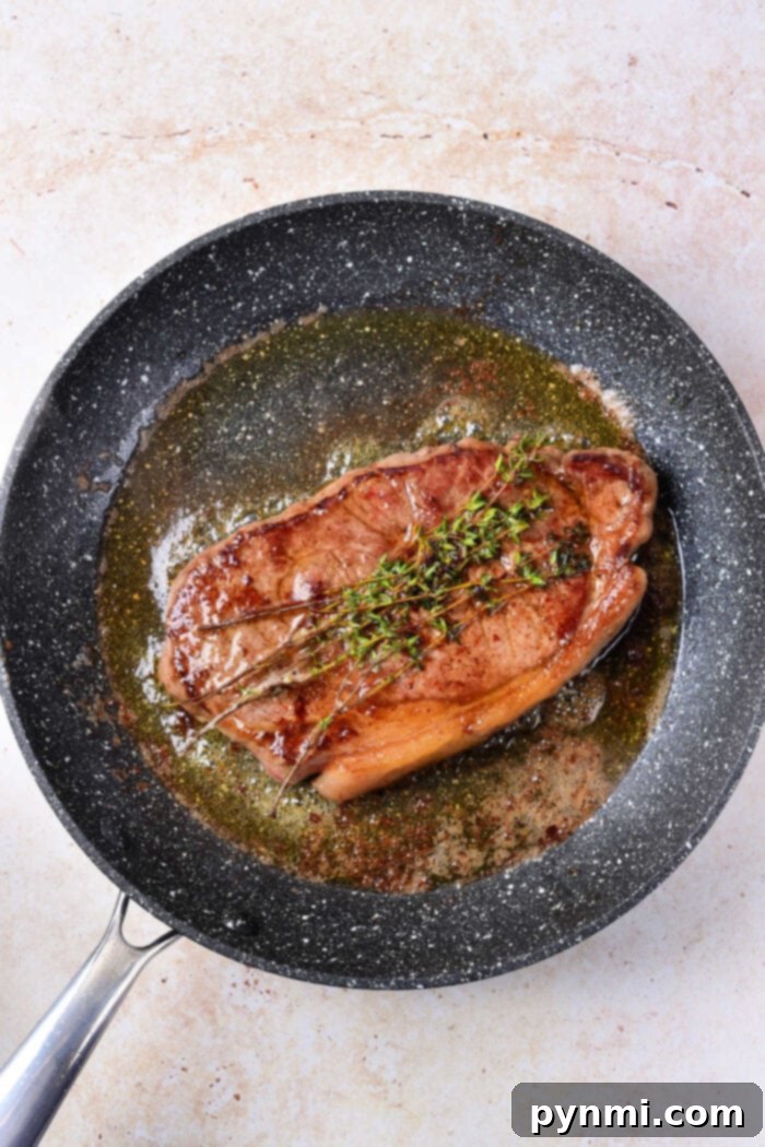 A perfectly cooked strip loin steak, garnished with fresh sprigs of thyme, rests in a black non-stick frying pan. The steak sits in a small pool of rendered fat and herbs, highlighting its rich, juicy appearance. This image showcases the final stage of cooking, just before resting and slicing.