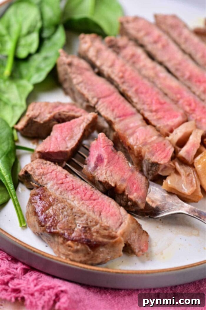 A plate showcasing perfectly sliced medium-rare steak, with prominent pink centers, demonstrating optimal doneness for beef strip loin steaks. The steak is neatly arranged next to a vibrant leafy green, possibly spinach or kale, adding a fresh element to the dish. A fork holds a piece of steak in the foreground, ready to be enjoyed, with a soft pink cloth providing a subtle background texture.