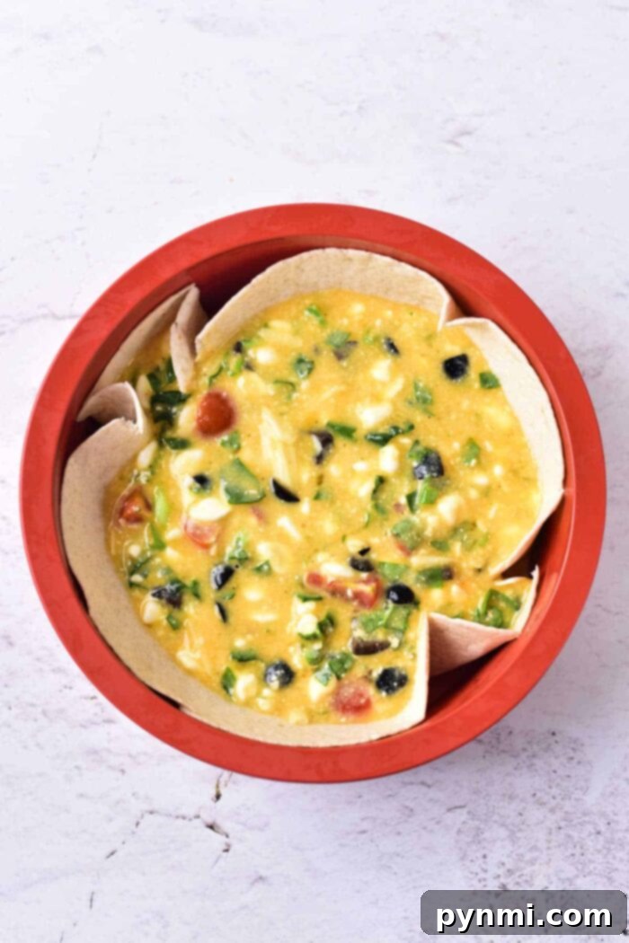 Light and Creamy Cottage Cheese Quiche 6 A vibrant red bowl showcasing a rich yellow cottage cheese quiche mixture, generously studded with colorful chopped vegetables such as olives, cherry tomatoes, green peppers, and corn. The bowl is neatly lined with pieces of tortilla, forming a makeshift crust. The background features a crisp, white textured surface.