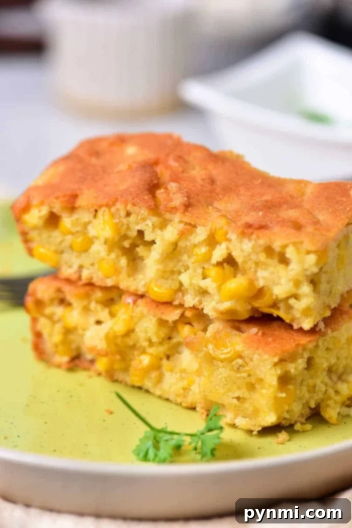 Two pieces of corn pudding with visible corn kernels stacked on a light green plate remind one of traditional Corn Pudding Without Jiffy Mix. A piece of parsley adorns the plate, and a fork is partially visible in the background.