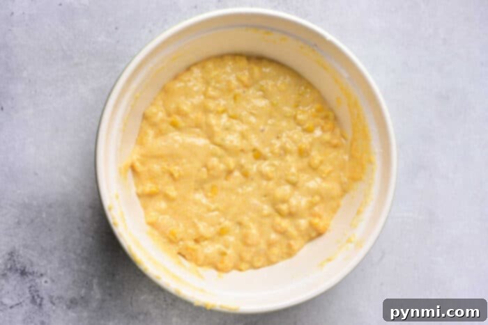 A white bowl filled with thick, yellowish batter containing visible corn kernels, placed on a light gray surface—perfect for homemade Corn Pudding Without Jiffy Mix.