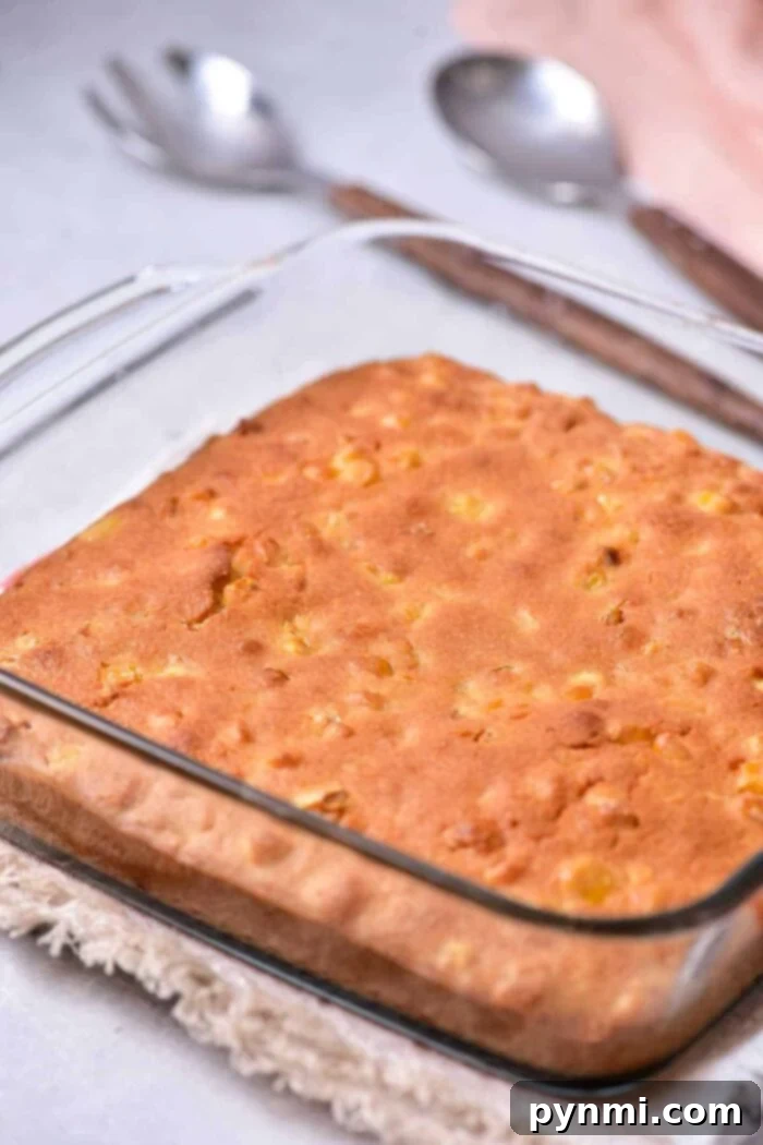 A square glass baking dish holds a freshly baked corn pudding with a golden-brown crust, reminiscent of homemade corn pudding without Jiffy mix. The dish is placed on a tan woven trivet, and two silver spoons are in the background on a light-colored surface.