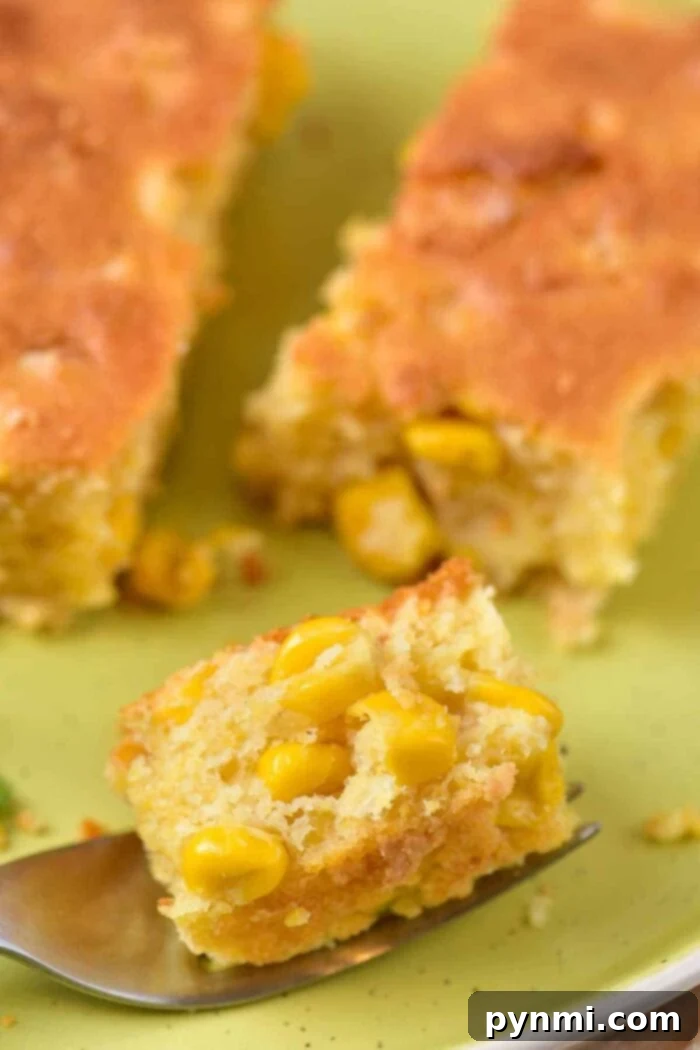 A close-up of a piece of corn casserole with corn kernels, reminiscent of corn pudding without Jiffy mix, placed on a green plate. A fork holding a smaller piece of the cornbread is visible in the foreground.