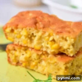 Two pieces of corn pudding with visible corn kernels stacked on a light green plate remind one of traditional Corn Pudding Without Jiffy Mix. A piece of parsley adorns the plate, and a fork is partially visible in the background.
