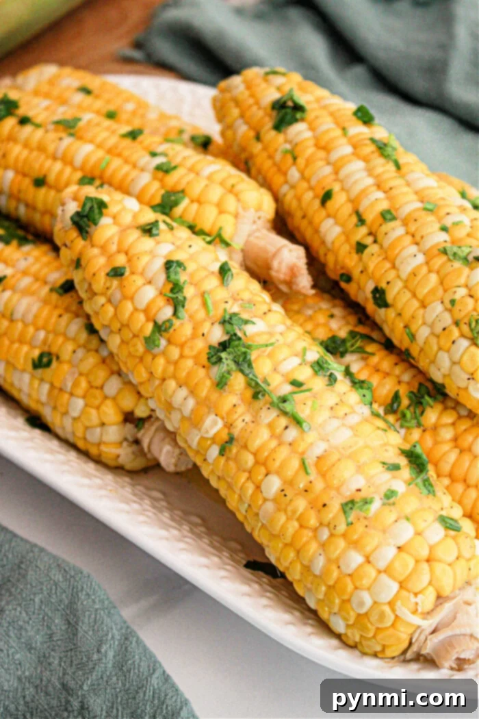 Tender Slow Cooker Corn Perfection 2 A close-up of several ears of Crockpot Corn on the Cob placed on a white rectangular dish. The corn is garnished with chopped fresh herbs and a sprinkle of seasoning. A green cloth is partially visible in the background.