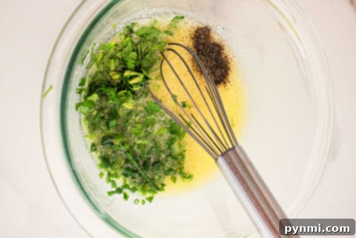 Tender Slow Cooker Corn Perfection 4 A glass mixing bowl containing a yellow liquid with finely chopped green herbs, ground black pepper, and a metal whisk, reminiscent of seasonings for Crockpot Corn on the Cob. The bowl is placed on a light-colored surface.
