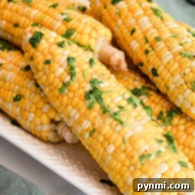 A close-up of several ears of Crockpot Corn on the Cob placed on a white rectangular dish. The corn is garnished with chopped fresh herbs and a sprinkle of seasoning. A green cloth is partially visible in the background.