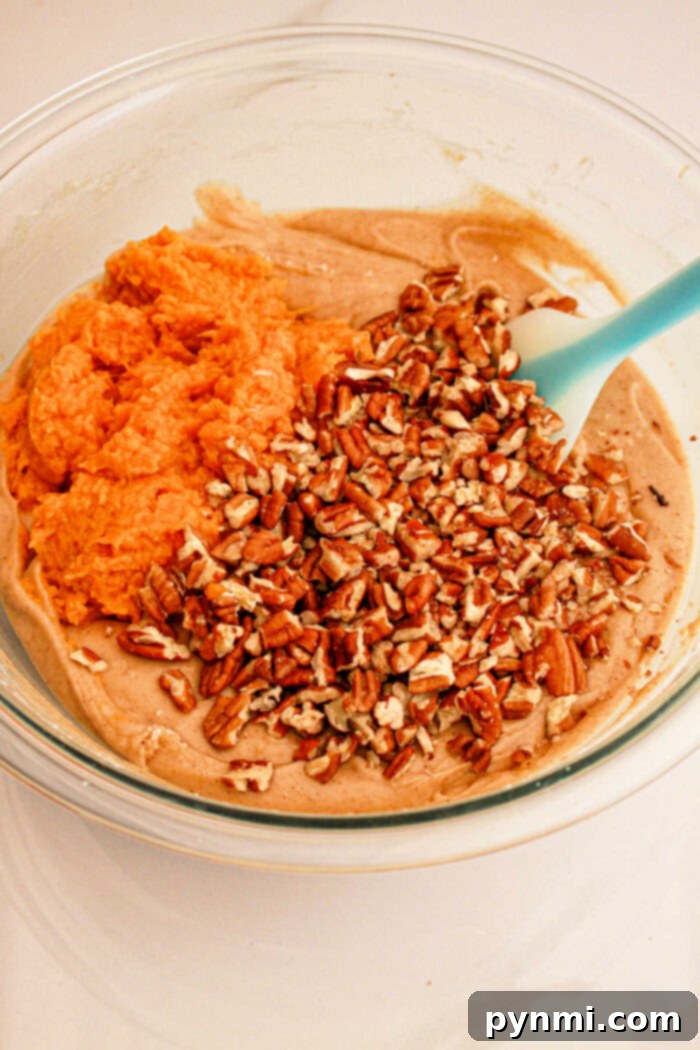 A glass bowl contains a mixture of ingredients being combined: creamy batter, orange sweet potato puree, and chopped pecans. A blue spatula is resting in the bowl, ready for mixing what will become delicious Sweet Potato Bread with Cake Mix.