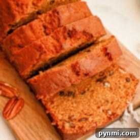 A loaf of sweet potato bread partially sliced on a wooden cutting board. The bread is golden brown with visible specks of nuts. Whole pecans and a cinnamon stick are placed beside the board on a white surface. A gray cloth is partially visible in the corner.