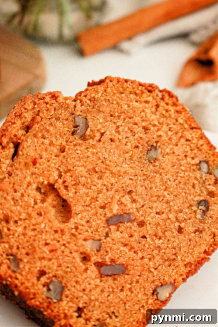 A close-up shot of a slice of pumpkin bread. The bread has a moist texture with visible pieces of nuts embedded throughout, reminiscent of sweet potato bread with cake mix. The background includes blurred elements hinting at additional ingredients or kitchen utensils.