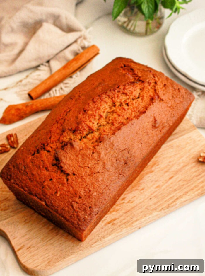 A loaf of freshly baked sweet potato bread with cake mix sits on a wooden cutting board. A cinnamon stick and some whole pecans are visible in the background, alongside a folded beige cloth and a stack of white plates.