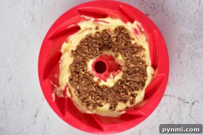 A bundt pan, prepped and ready, holds a layer of yellow cake batter, topped with the cinnamon-pecan crumble, waiting for the final layer of batter to create the classic Sock It To Me Cake swirl.