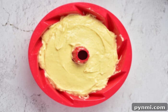 A vibrant red bundt pan, glistening with non-stick spray, holds a perfectly smooth layer of golden yellow cake batter, ready for its next step in becoming a delicious Sock It To Me Cake.