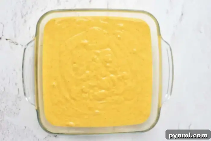 Pumpkin Perfection with Yellow Cake Mix 4 A square glass dish filled with a smooth, yellow batter sits on a light-colored marbled surface. The batter, perfect for pumpkin cake with yellow cake mix, has a few small bubbles scattered across the top, indicating that it has just been prepared and is ready to be baked.