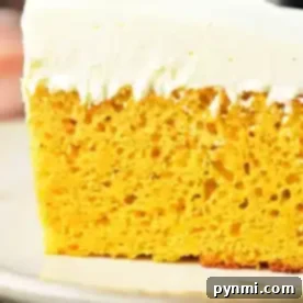 A close-up of a slice of pumpkin cake with yellow cake mix, featuring a thick layer of white frosting. The cake has a yellow, airy interior with visible bubbles and is placed on a beige plate.