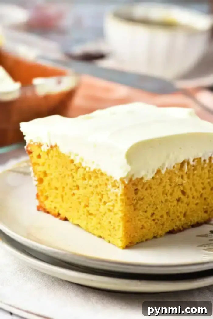 Pumpkin Perfection with Yellow Cake Mix 7 A close-up image of a slice of yellow pumpkin cake with a thick layer of white frosting on a plate. The background includes a blurred view of more frosted cake and tableware, including a knife and a bowl. The scene is set on a light-colored tablecloth.