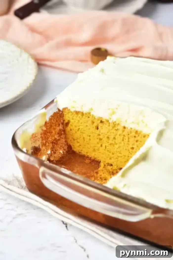 Pumpkin Perfection with Yellow Cake Mix 8 A rectangular glass baking dish containing a pumpkin cake with yellow cake mix, topped with white frosting. A square piece has been removed, revealing the cake's interior. The dish sits on a light-colored cloth, with a plate and peach-colored fabric in the background.