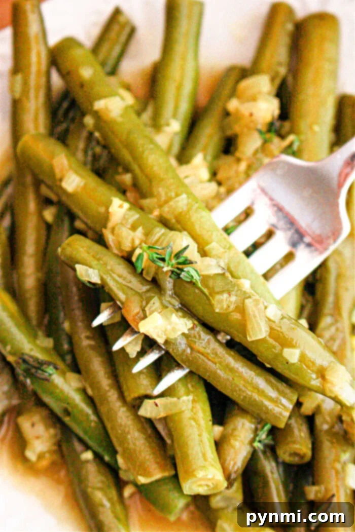 Tender Slow Cooker Green Beans 2 A close-up image of crockpot-cooked green beans seasoned with chopped onions and herbs. A fork is piercing several of the green beans, and the dish appears to be served on a white plate.