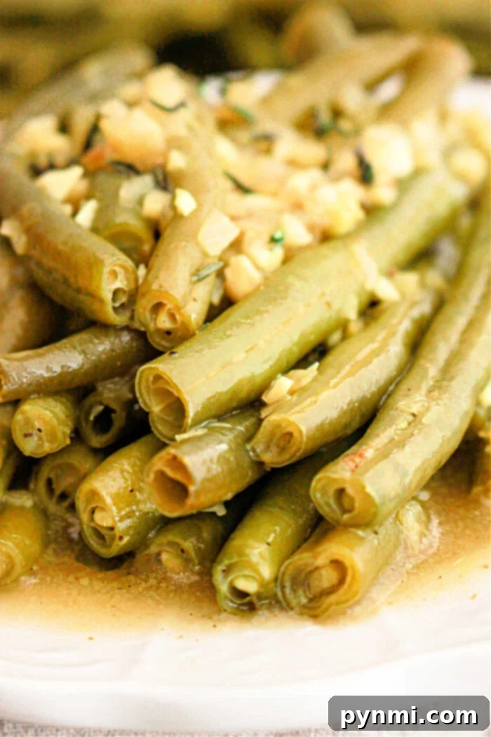Tender Slow Cooker Green Beans 6 A close-up of crockpot-cooked green beans topped with finely chopped garlic and herbs. The green beans are slightly glossy from the seasoning and arranged in a pile on a white plate.