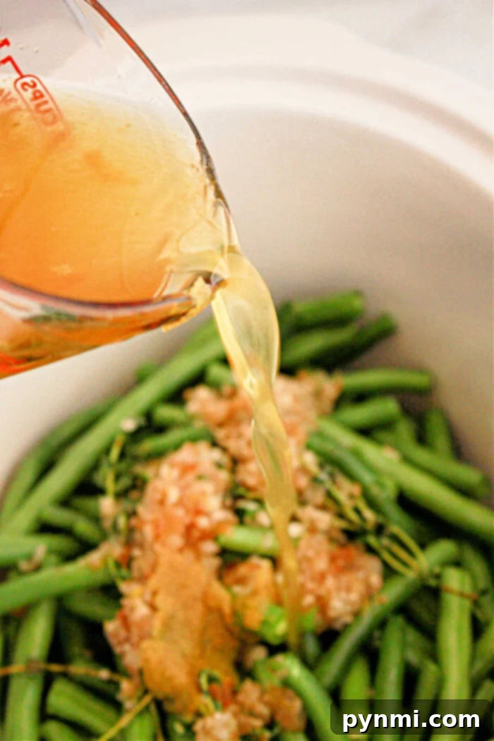 Tender Slow Cooker Green Beans 8 Close-up of a measuring cup pouring broth into a crockpot filled with green beans, minced garlic, and other seasonings. The green beans are uncooked, and the garlic appears to be spread on top.