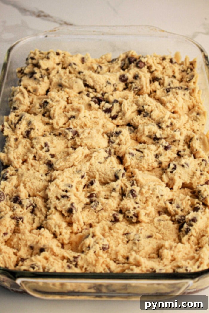 Ultimate Brownie Cookie Dough Brookie Bars 4 Close-up of a clear glass baking dish evenly spread with unbaked chocolate chip cookie dough. The light brown dough, studded with visible chocolate chips, is perfectly layered atop a brownie base, illustrating the easy second step in preparing delicious brookies with brownie mix and cookie dough before baking.