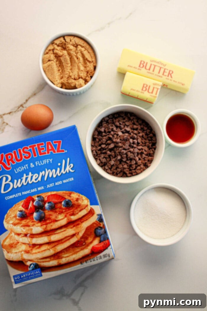 Ingredients for a baking recipe are laid out on a counter: a box of pancake mix perfect for cookies, a bowl of brown sugar, chocolate chips, two sticks of butter, vanilla extract in a small cup, white sugar in another bowl, and one egg.
