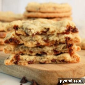 A stack of chocolate chip cookies made with pancake mix is halved and placed on a wooden board. The cookies are thick with visible chunks of melted chocolate. Some chocolate chips are scattered around the board, and more pancake mix cookies are stacked in the background.