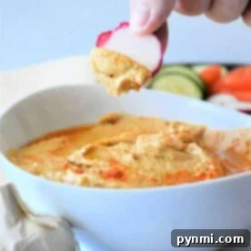 Hand dipping sliced radish into a creamy, easy hummus made with canned chickpeas, with garlic and fresh vegetables in the background.
