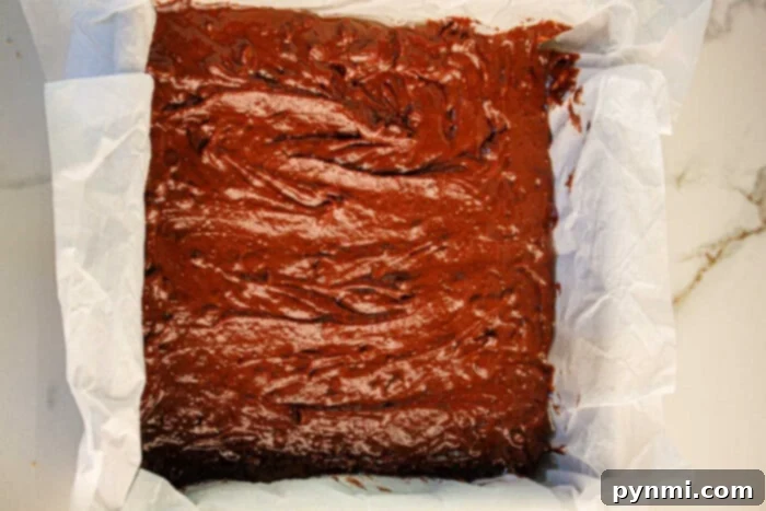 Secret Ingredient Chocolate Black Bean Brownies 5 A parchment-lined square baking tin rests on a marble surface, brimming with rich chocolate black bean brownie batter, ready to mix magic in the oven.
