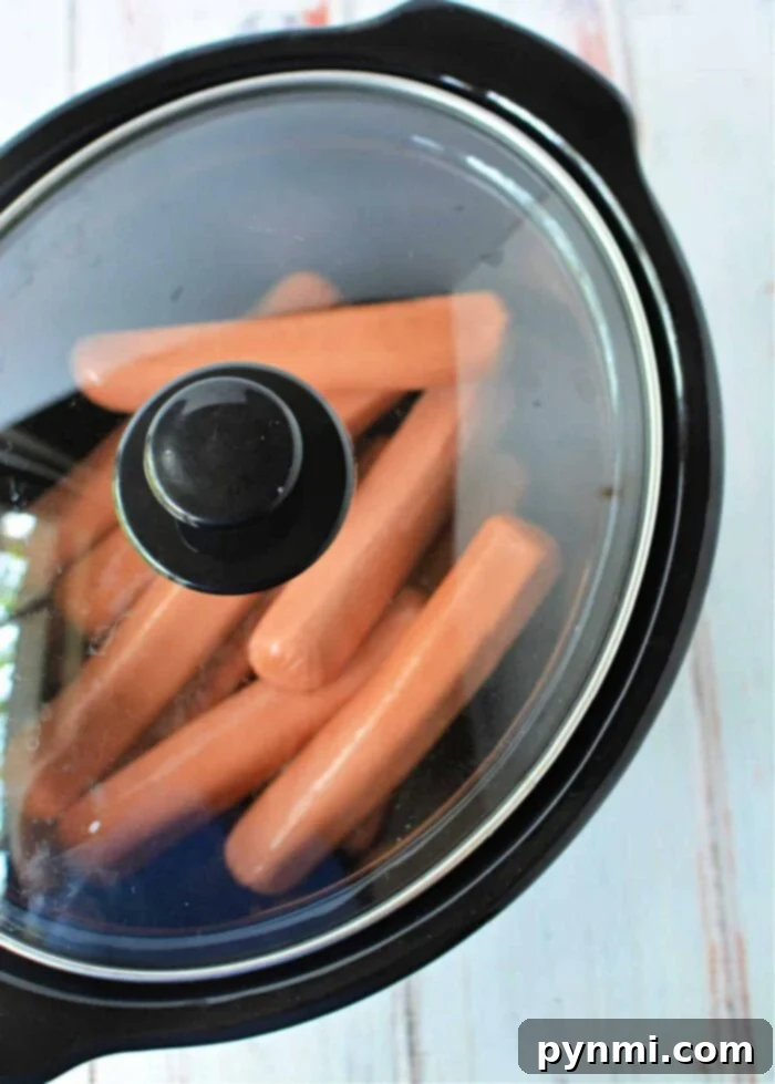 Speedy Slow Cooker Hot Dogs 2 A sleek black slow cooker with its glass lid removed, revealing a generous batch of perfectly cooked, plump hot dogs nestled inside. The appliance is placed on a subtly patterned white surface, ready to serve a large gathering.