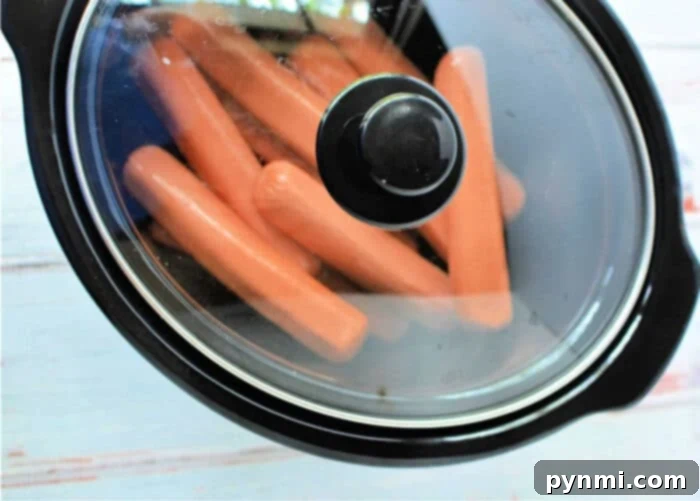 Speedy Slow Cooker Hot Dogs 5 An inviting top-down view of a black slow cooker, its interior filled with an abundant arrangement of hot dogs. The glass lid partially covers the pot, allowing a glimpse of the steaming wieners, all set on a light-colored surface.