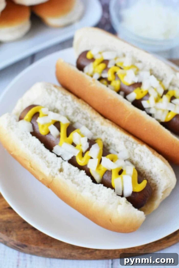 Speedy Slow Cooker Hot Dogs 7 Two perfectly prepared hot dogs in soft buns, artfully arranged on a clean white plate. The hot dogs, which were slow-cooked to tenderness, are generously garnished with finely diced onions and a classic swirl of yellow mustard. In the blurred background, a larger platter of slow cooker hot dogs and a bowl of fresh chopped onions indicate a ready spread for a party.