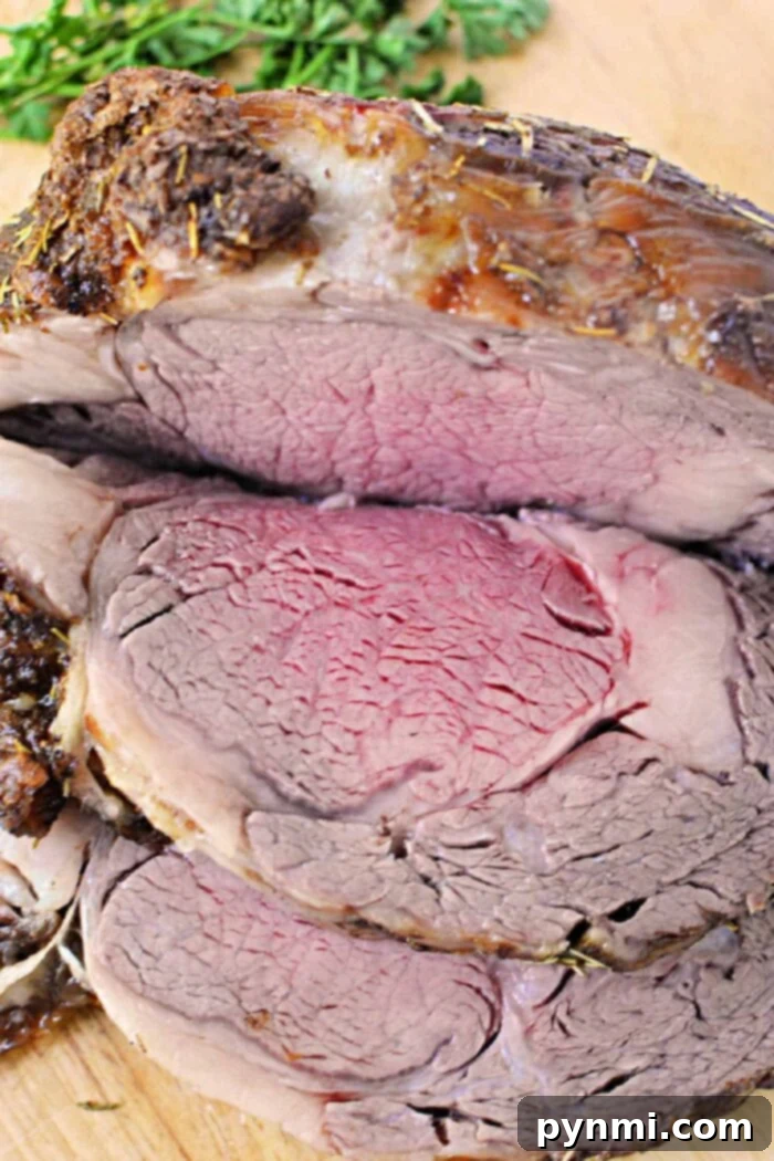 A close-up of a sliced, medium-rare Boneless Rib Roast. The beef is browned on the outside and pink with a slightly red center on the inside. It is garnished with sprigs of fresh herbs in the background. The meat is placed on a wooden cutting board, ready to be served.
