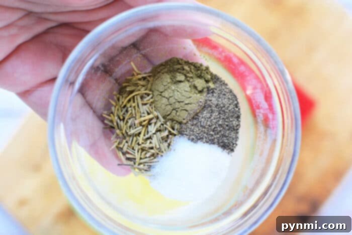 A hand holding a clear glass bowl containing a mix of dried rosemary, ground black pepper, salt, and another greenish-brown spice. This simple yet effective dry rub is perfect for seasoning your Boneless Rib Roast Recipe, infusing it with aromatic flavors.