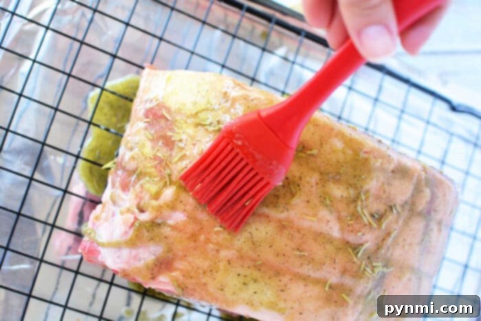 A close-up of a boneless rib roast on a metal cooling rack, covered in foil, being brushed with seasoning using a red silicone basting brush. The meat has a coat of a yellowish marinade with visible herbs and spices. A hand is holding the basting brush, ensuring every part of the rib roast recipe is infused with flavor.